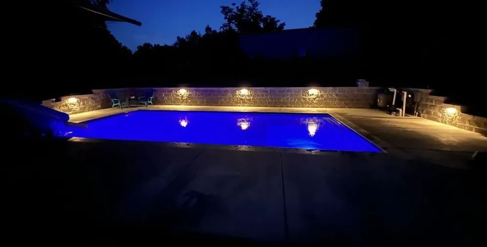 Nighttime view of a rectangular pool with bright blue water, lit by surrounding lights. Dark setting.