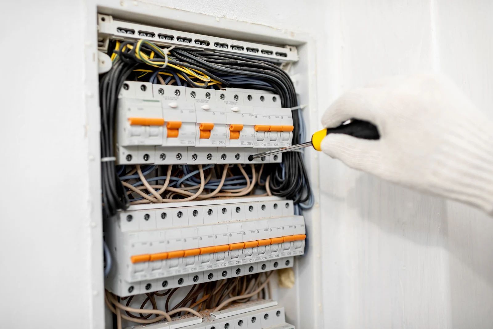 Person in white glove using screwdriver on a circuit breaker panel with various wires.