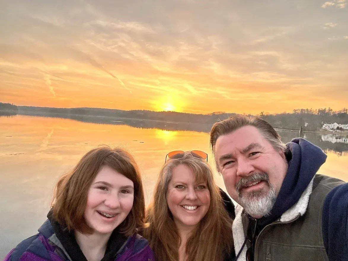 A man and two women are posing for a picture in front of a lake at sunset.