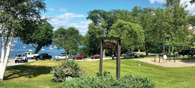 A park with a sign in the middle of it and cars parked in the background.