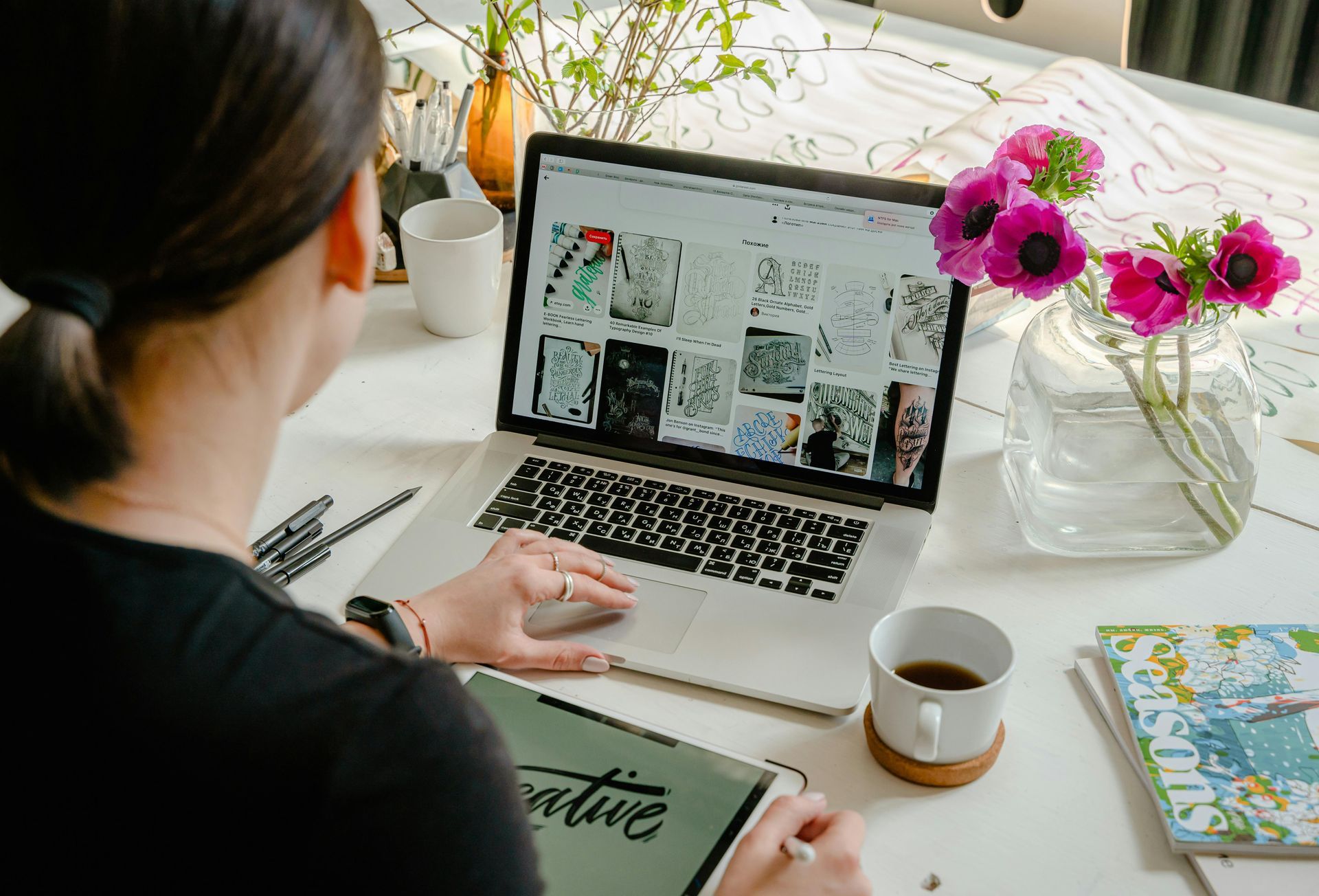 A person using a laptop at a desk with a vase of pink flowers and a mug nearby, browsing a grid of artistic designs.