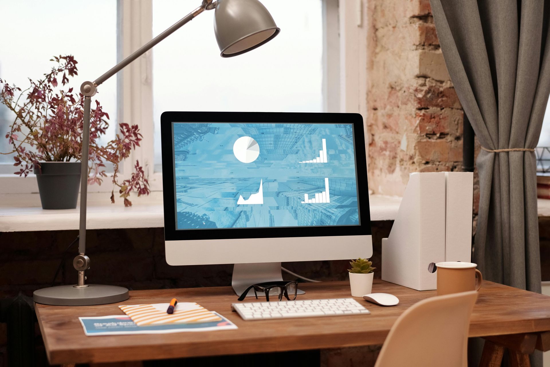 A computer monitor displays business charts on a desk in a home office with a lamp, plants, and a window.