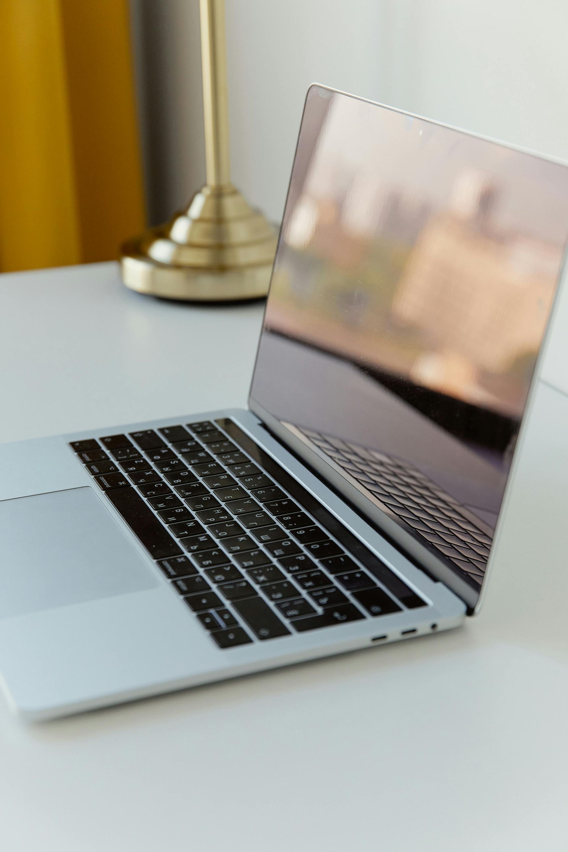 A silver laptop sits open on a clean, white desk with a brass lamp in the blurred background.