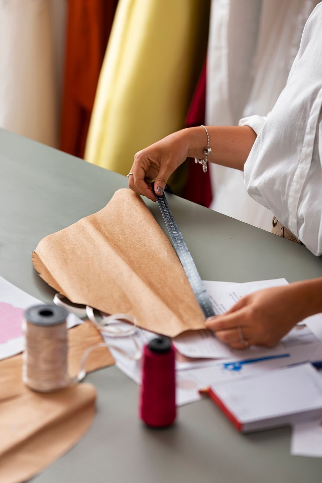 Una persona con camisa blanca mide una tela color canela con una regla en un escritorio, rodeada de carretes de costura y patrones.