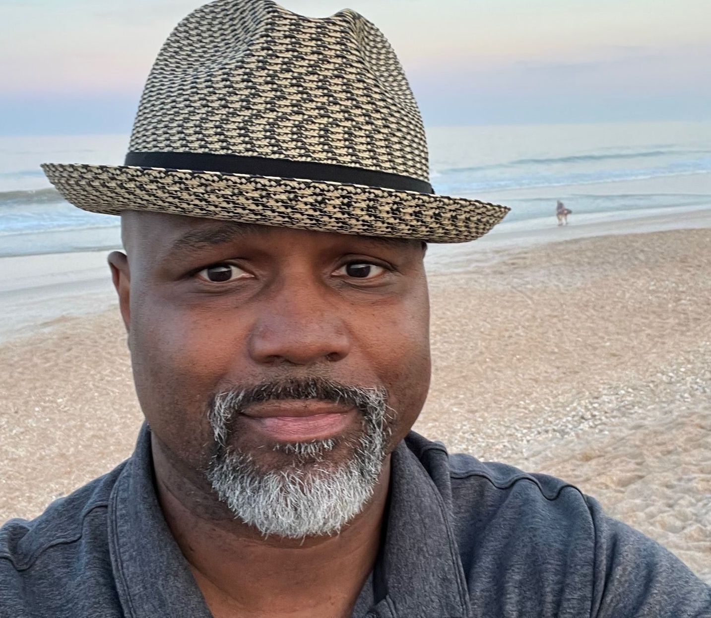 Man with a graying beard and hat smiles at the camera on a beach.