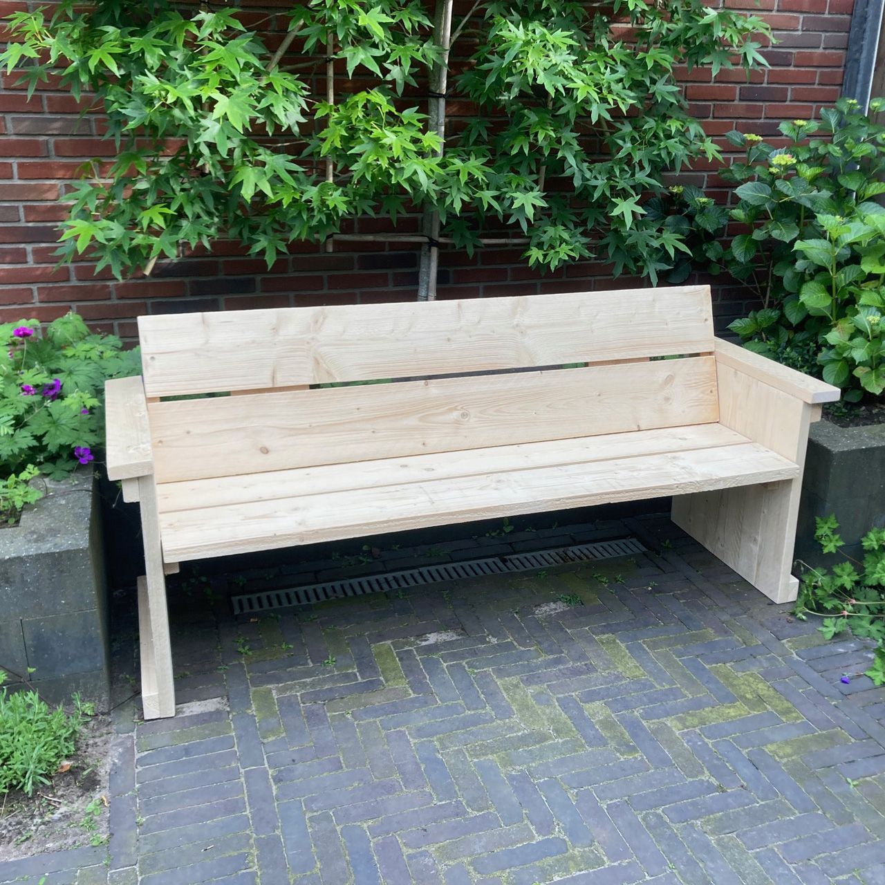 Steigerhouten tuinbank met armleuningen en rugleuning, geplaatst op een terras met klinkers, tegen een bakstenen muur met begroeiing