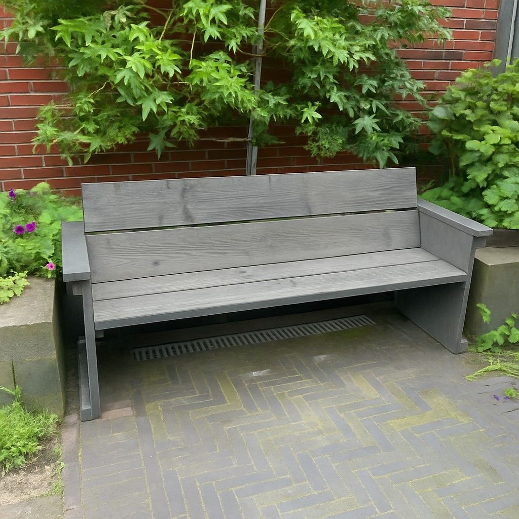 Steigerhouten tuinbank met armleuningen en rugleuning, geplaatst op een terras met klinkers, tegen een bakstenen muur met begroeiing