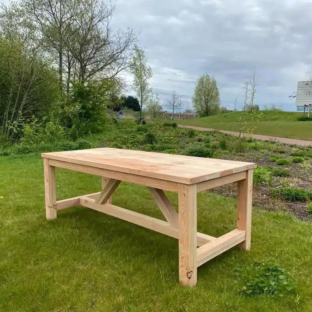 Houten tafel op het gras in een tuin, met bomen en een bewolkte hemel op de achtergrond.