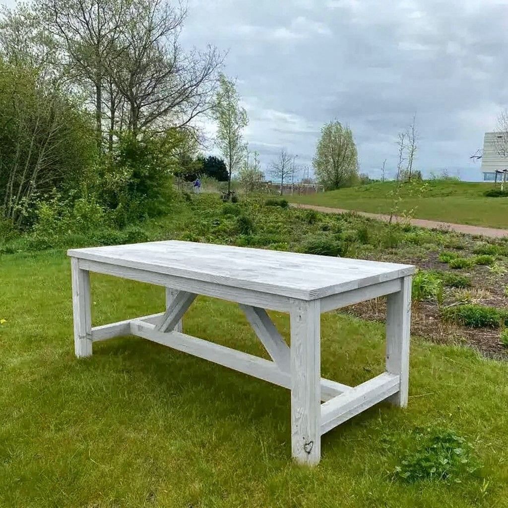 Witte houten tuintafel op een grasveld met bomen en een bewolkte hemel op de achtergrond.
