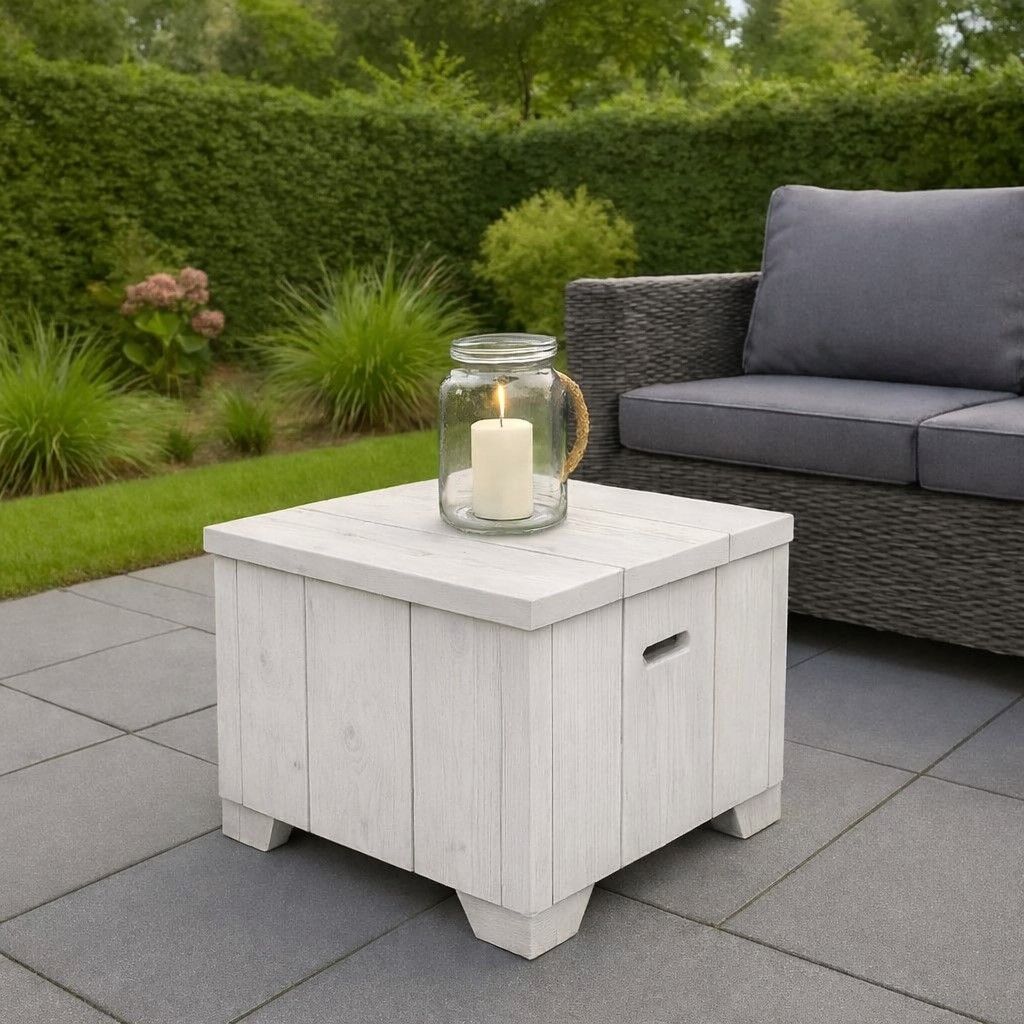 Witte opbergtafel voor buiten met een lantaarn op een grijs terras naast een rieten bank en een groene haag.