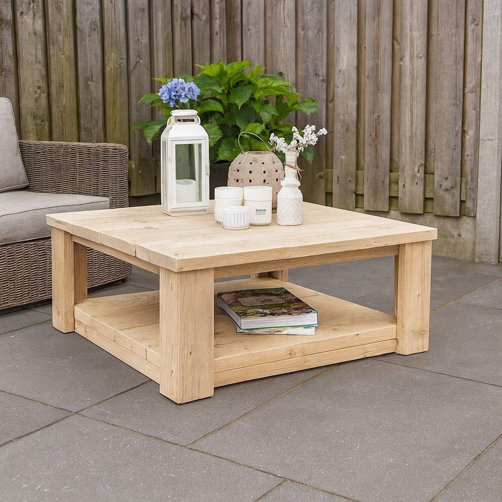 Een houten salontafel voor buiten met een lantaarn, bloemen, kaarsen en een boek op een terras naast een rieten bank.