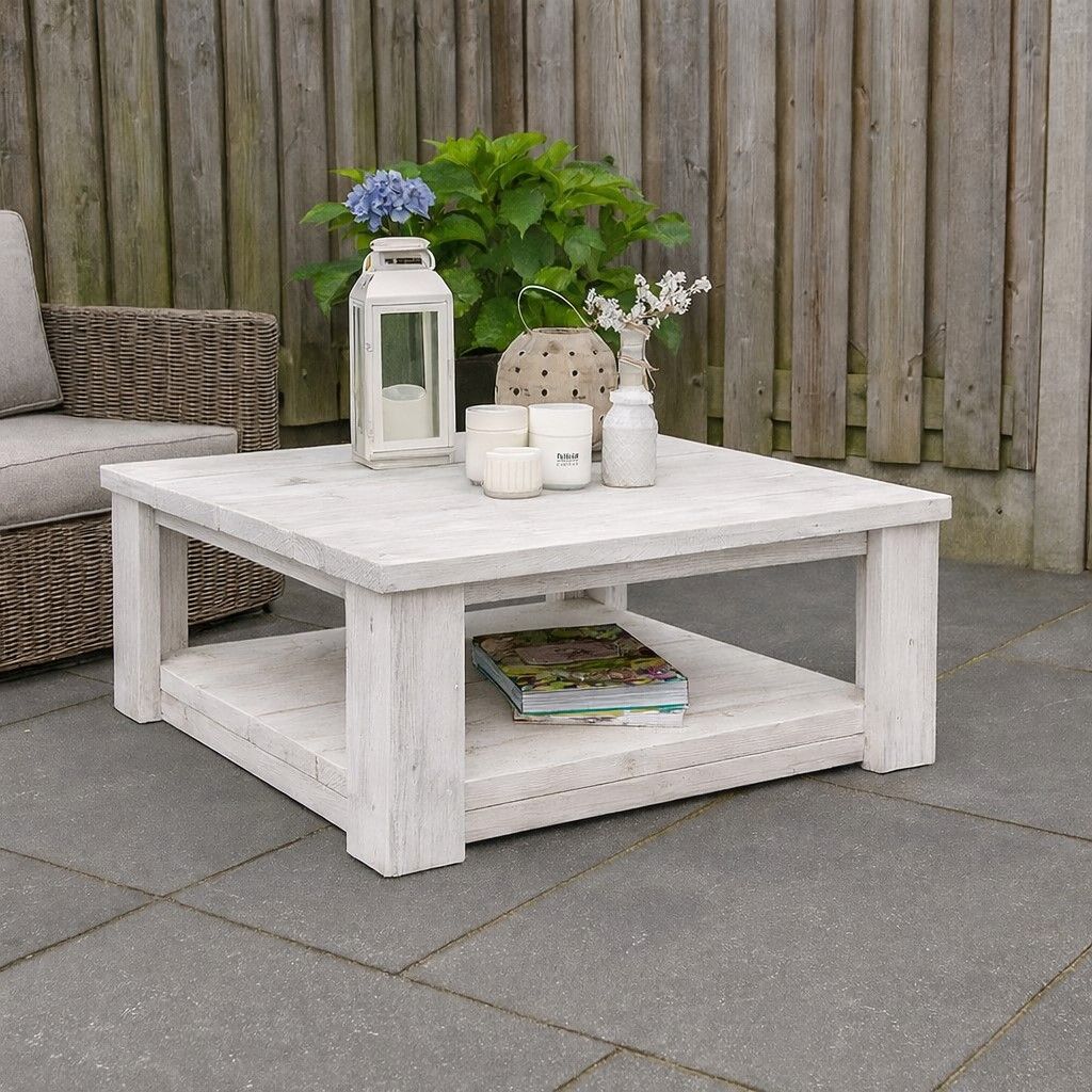 Witte salontafel voor buiten met lantaarn, kaarsen, bloemen en tijdschriften op een terras tegen een houten schutting.