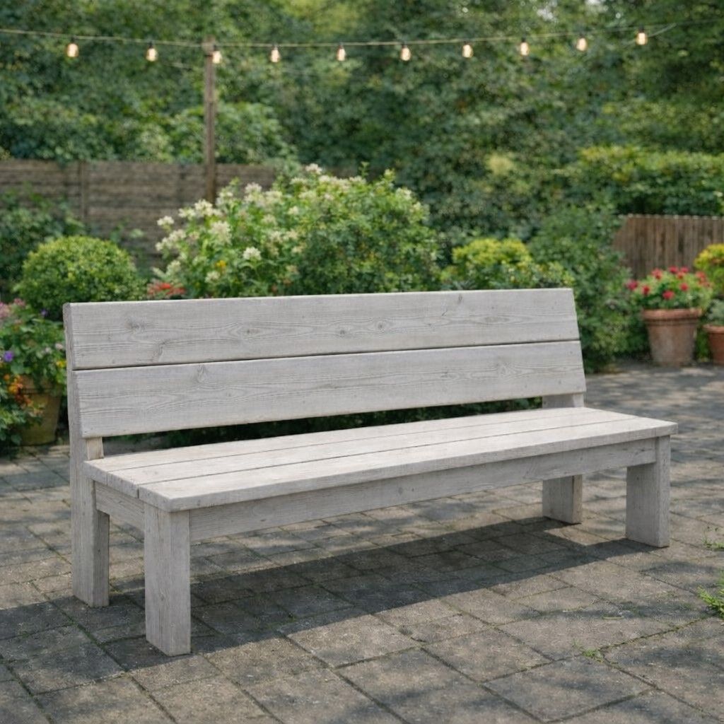 Een verweerde grijze houten bank in een binnentuin met struiken, potplanten en lichtslingers.