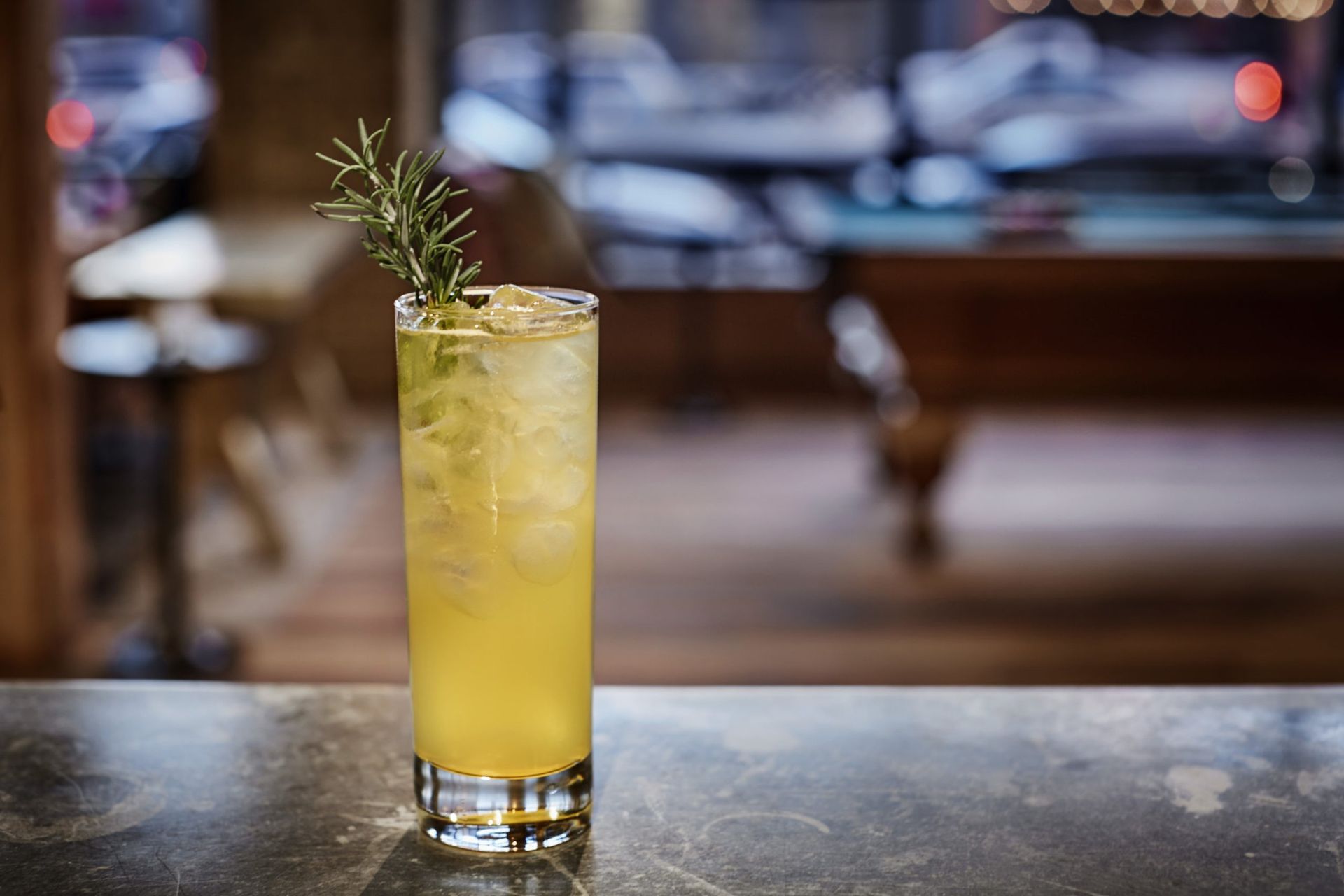 a tall glass filled with a yellow liquid and a sprig of rosemary on a table