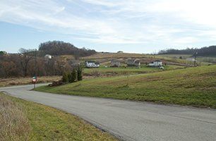 A road going through a grassy field with houses in the background.