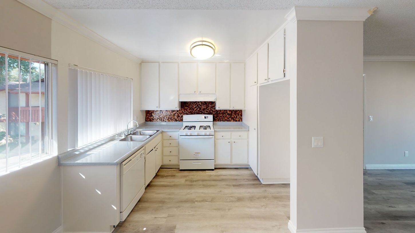Kitchen with white cabinets, appliances, and countertops. Reddish-brown backsplash, window with blinds, and light wooden floor.