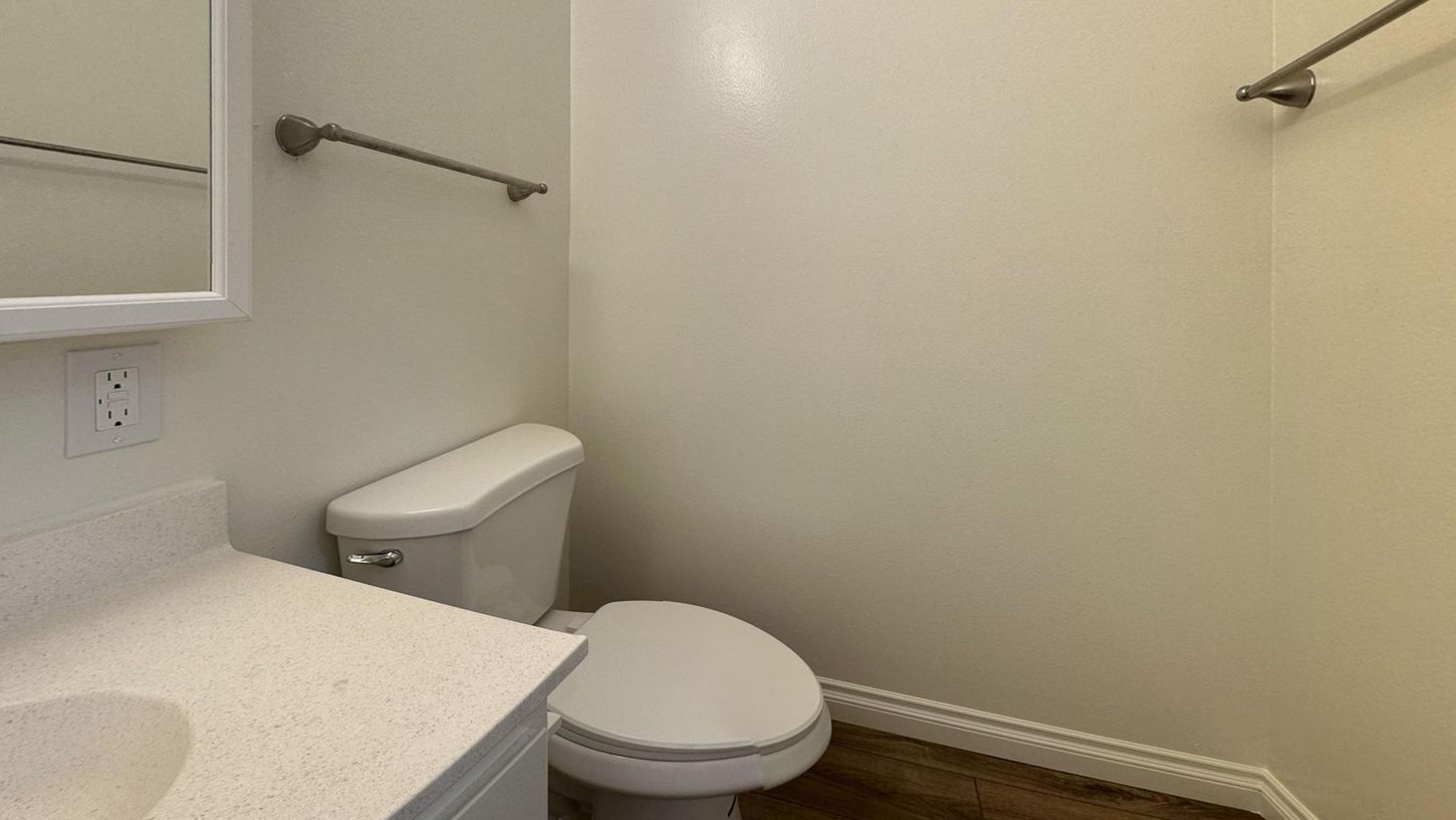 Bathroom with toilet, sink, and towel rack. White walls, wood-look floor, and a mirror are visible.