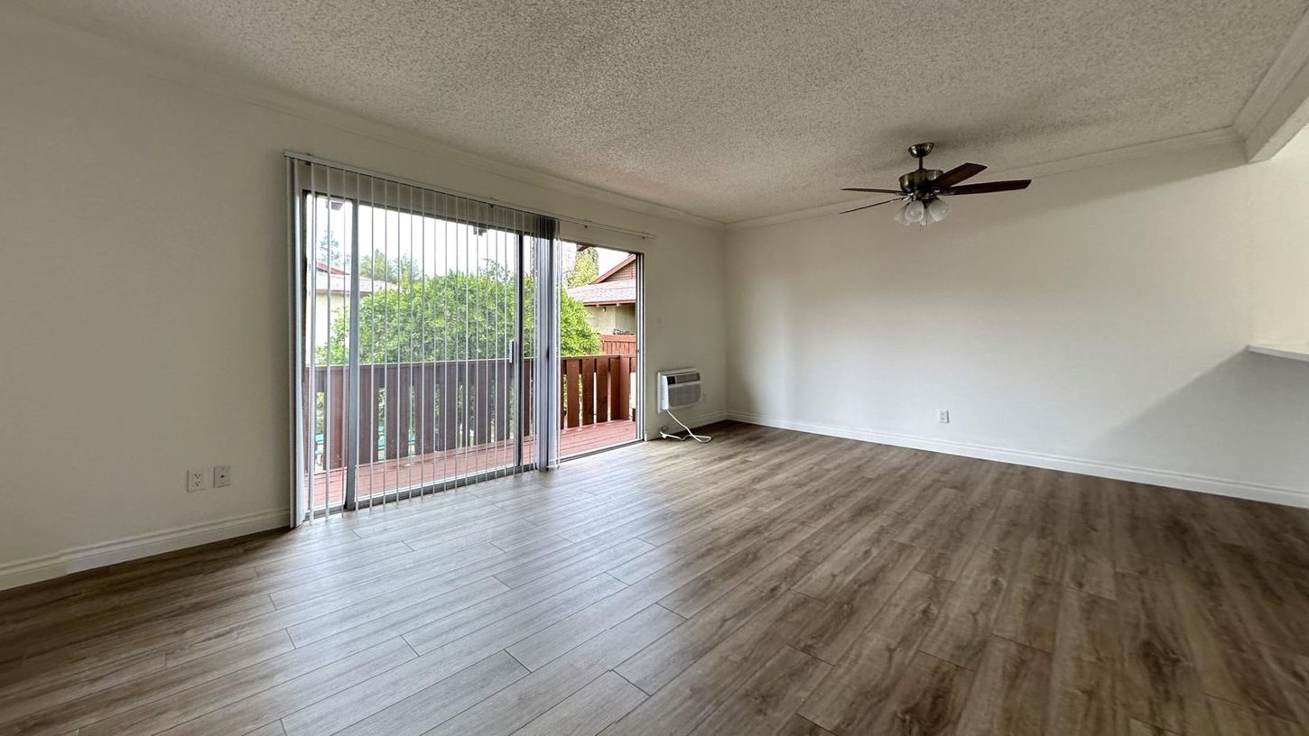 Empty room with sliding glass door, balcony, and ceiling fan; brown wood-look flooring.