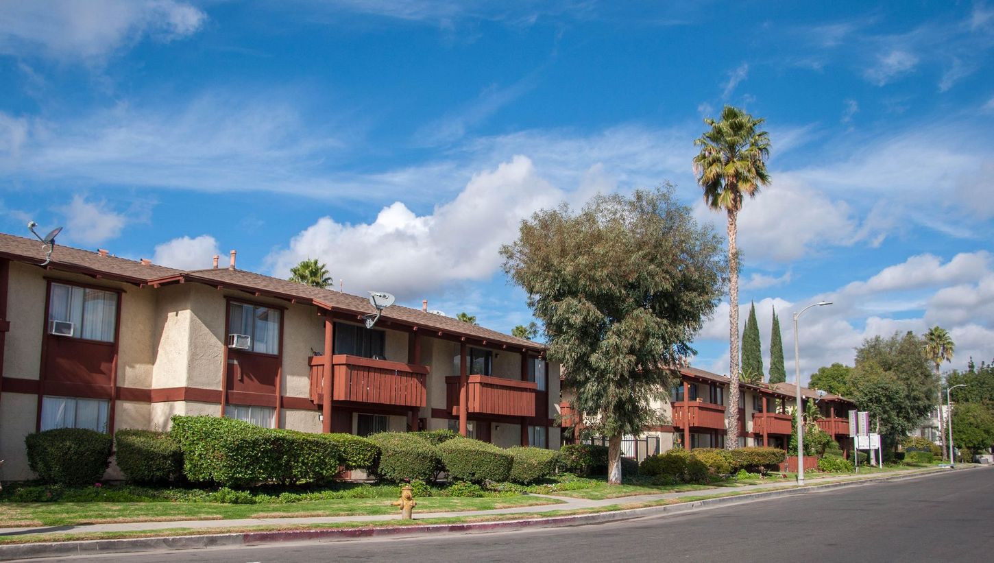 Two-story apartment complex with brown trim and balconies on a sunny day. Palm tree and cloudy blue sky.