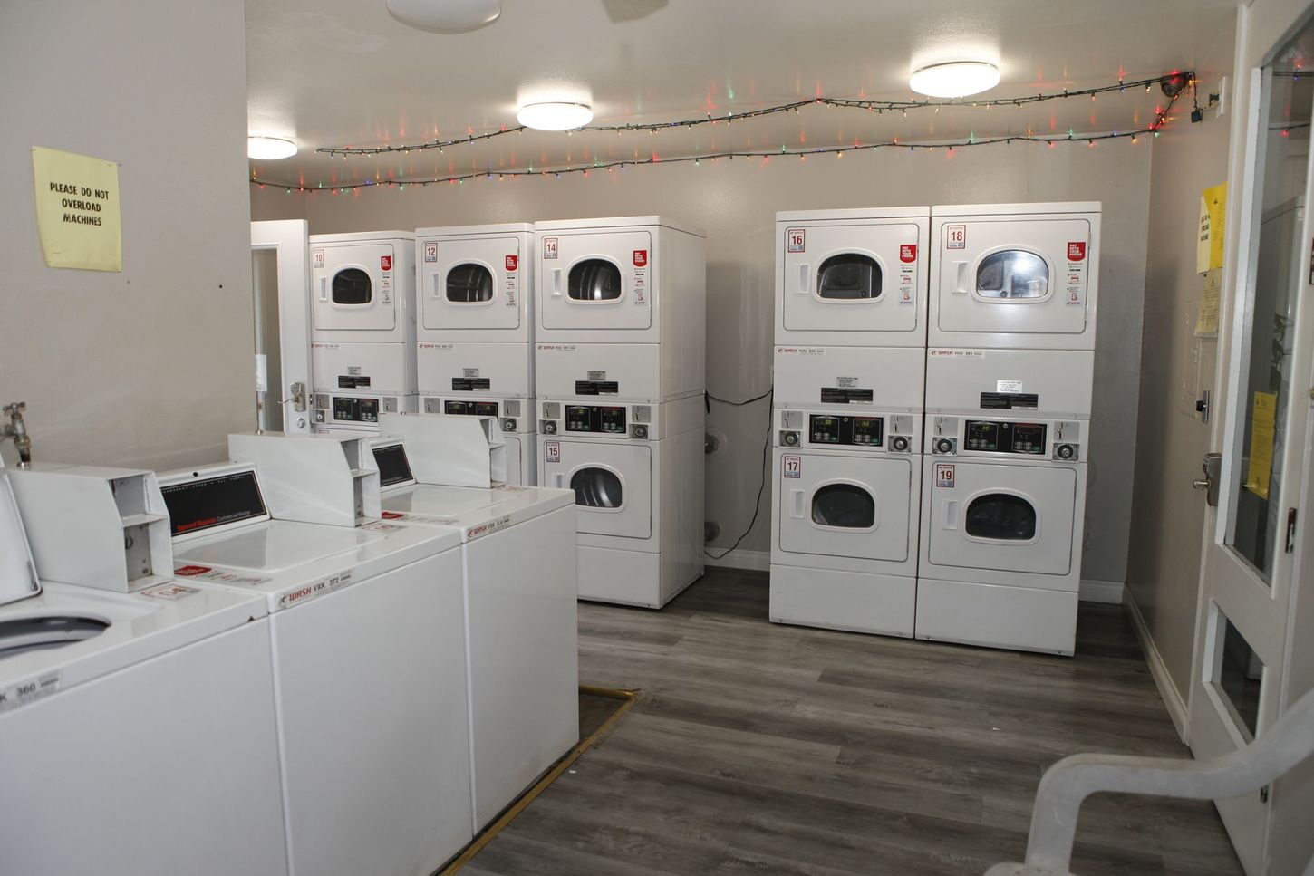 Laundry room with several white washers and dryers. Gray flooring and bright overhead lights.