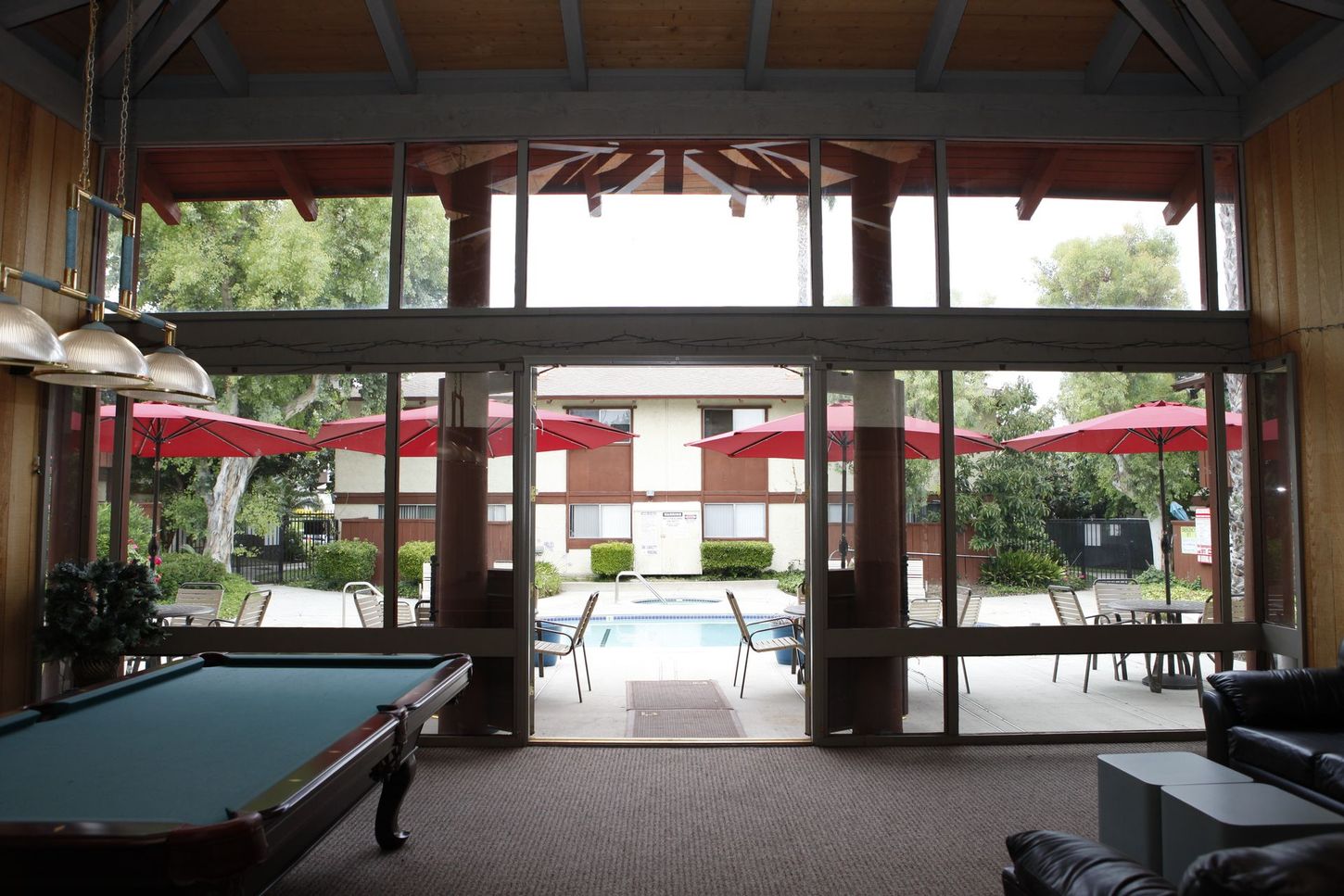 Pool room interior with a pool table, and a view of a pool area with red umbrellas and buildings.