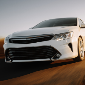White sedan driving on a road. The car is angled towards the viewer, with a blurred background. | RX Automotive