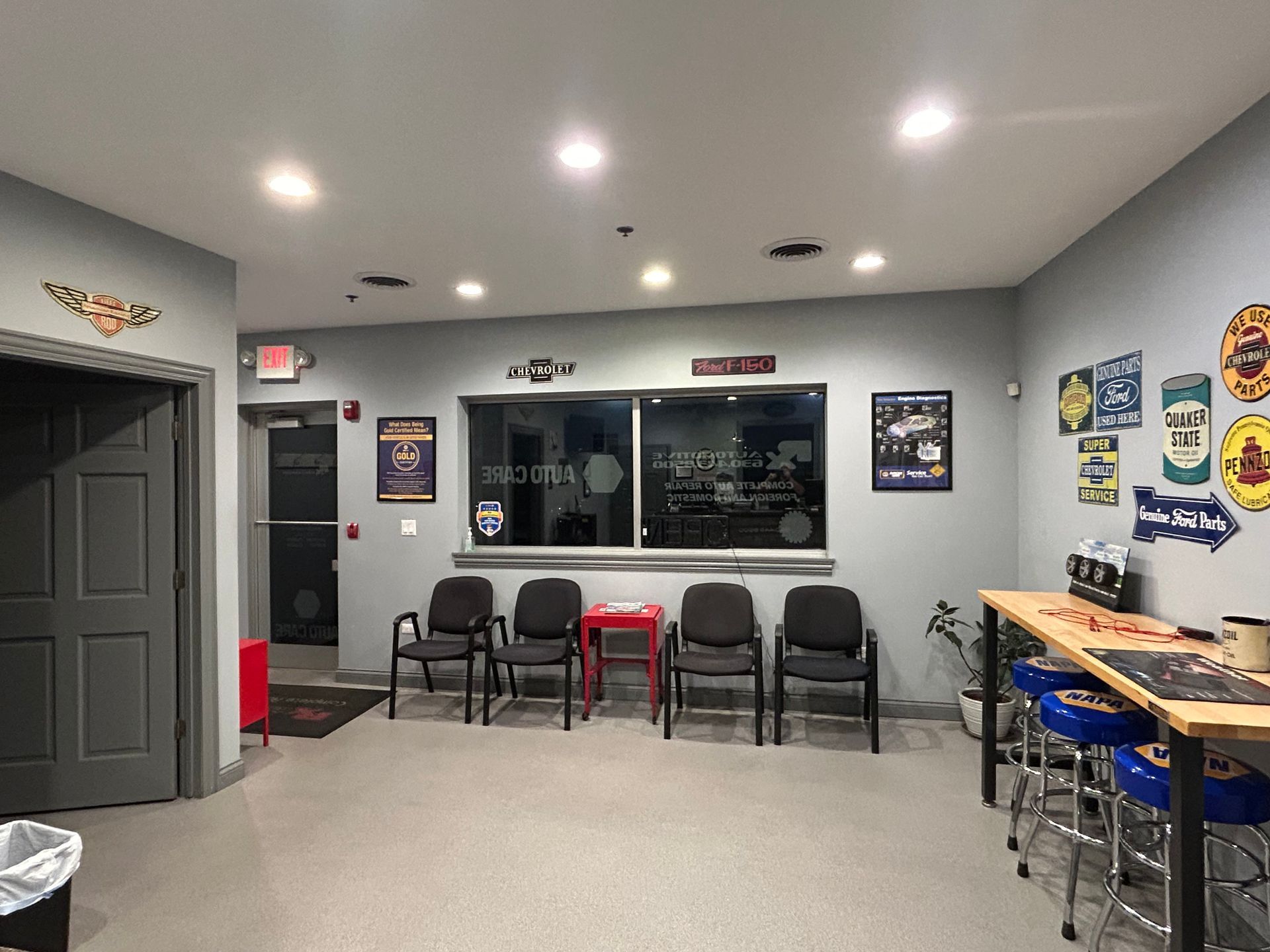 Waiting room with chairs, signs, and a bar-height table. Gray walls, a door on the left, and a window. | RX Automotive