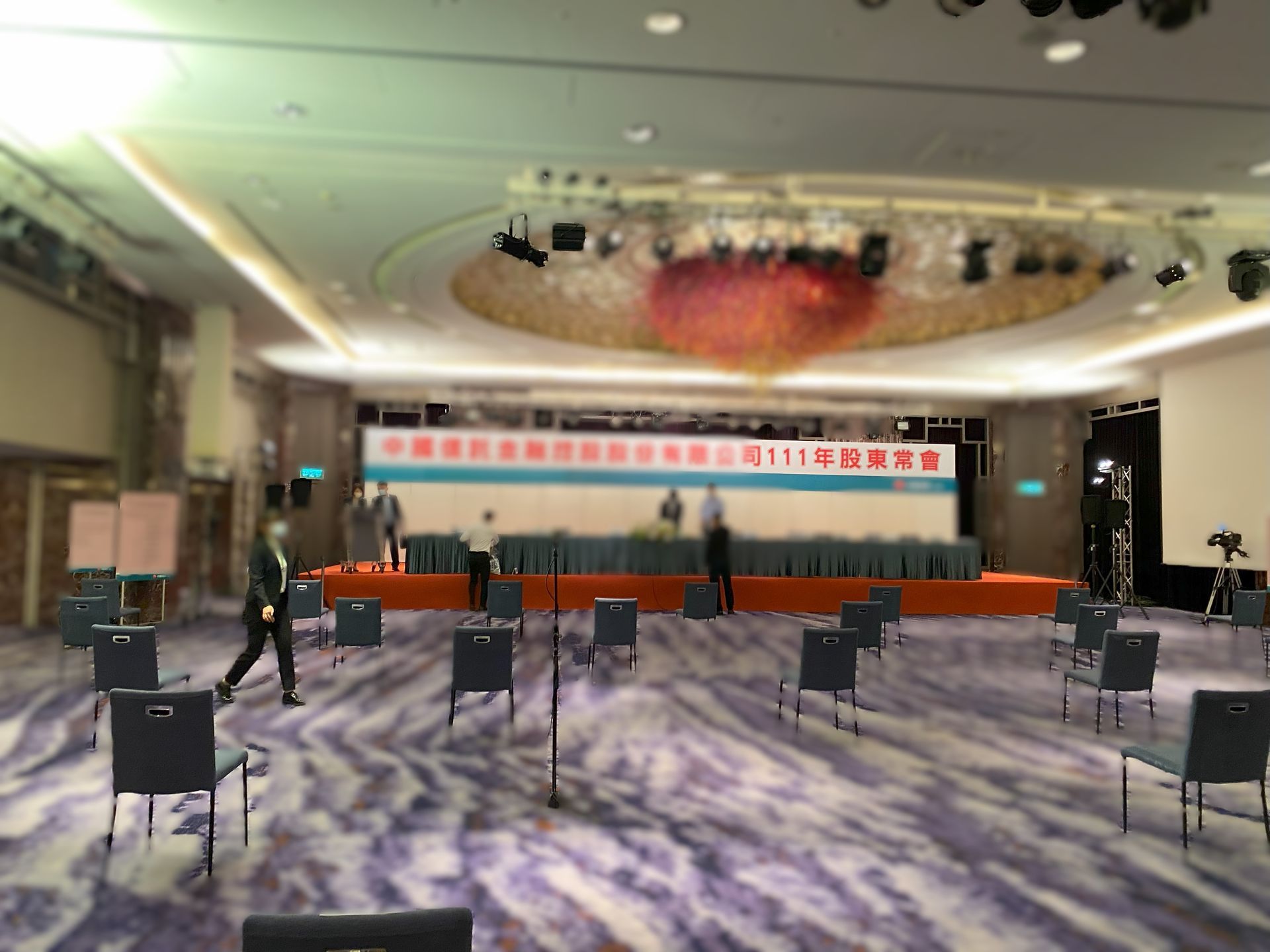 為活動佈置的大廳。椅子之間間隔開。可以看到舞台上坐著人。