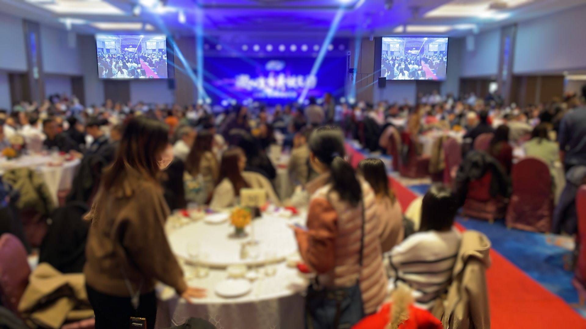 一個寬敞、模糊的宴會廳，裡面坐著客人，還有一個有螢幕的舞台；一場公司活動。