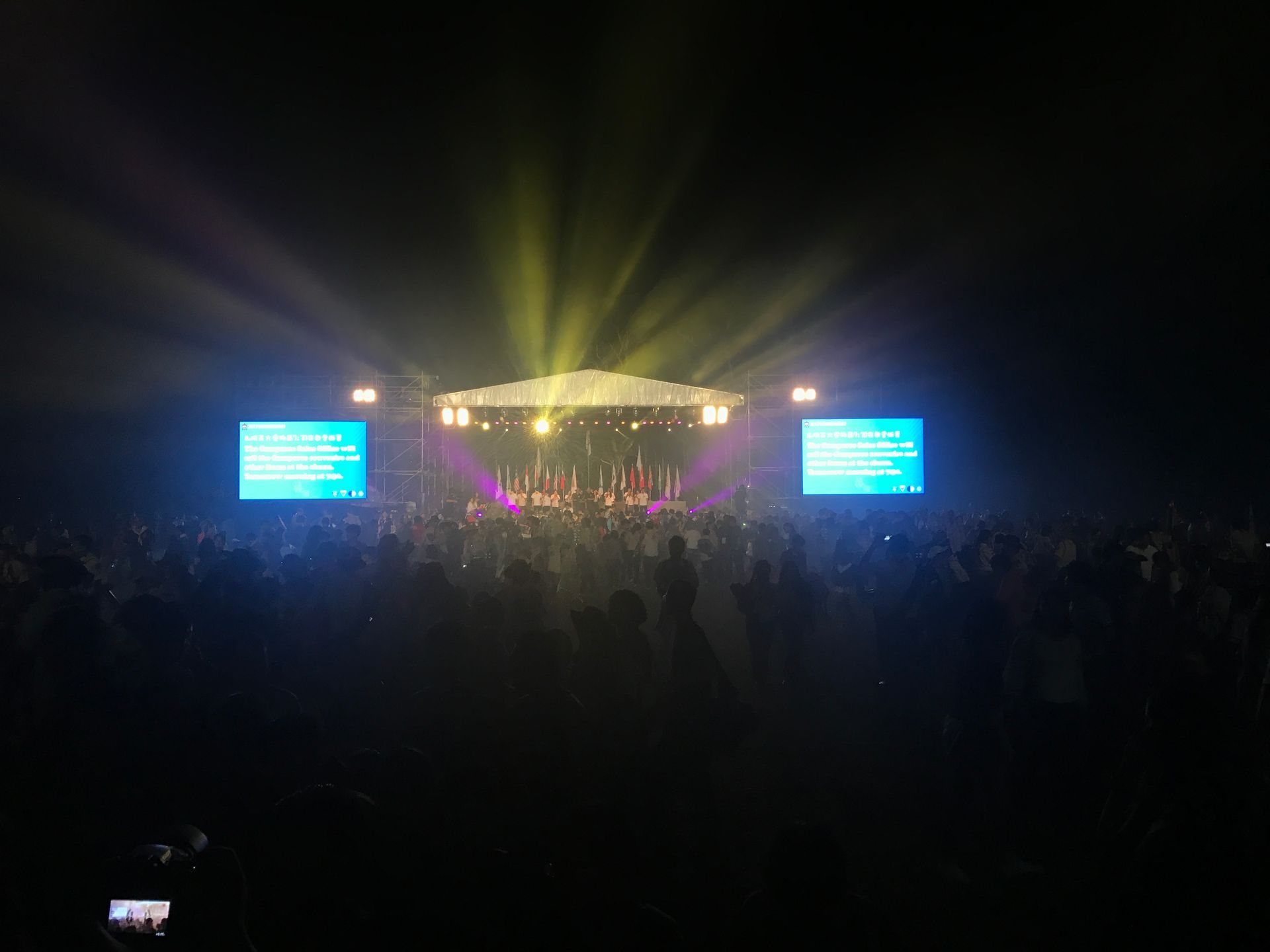 音樂會舞台被聚光燈照亮，人群處於黑暗中，兩個螢幕顯示文字。