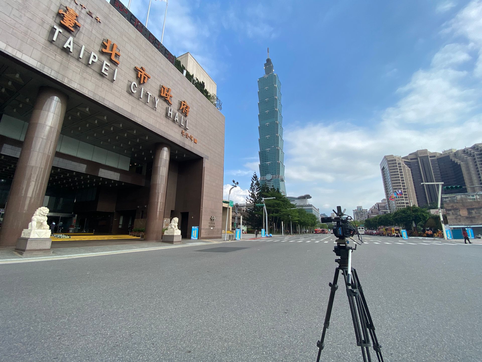 藍天下的台北市政廳和台北 101 大樓，前面有一個帶相機的三腳架。