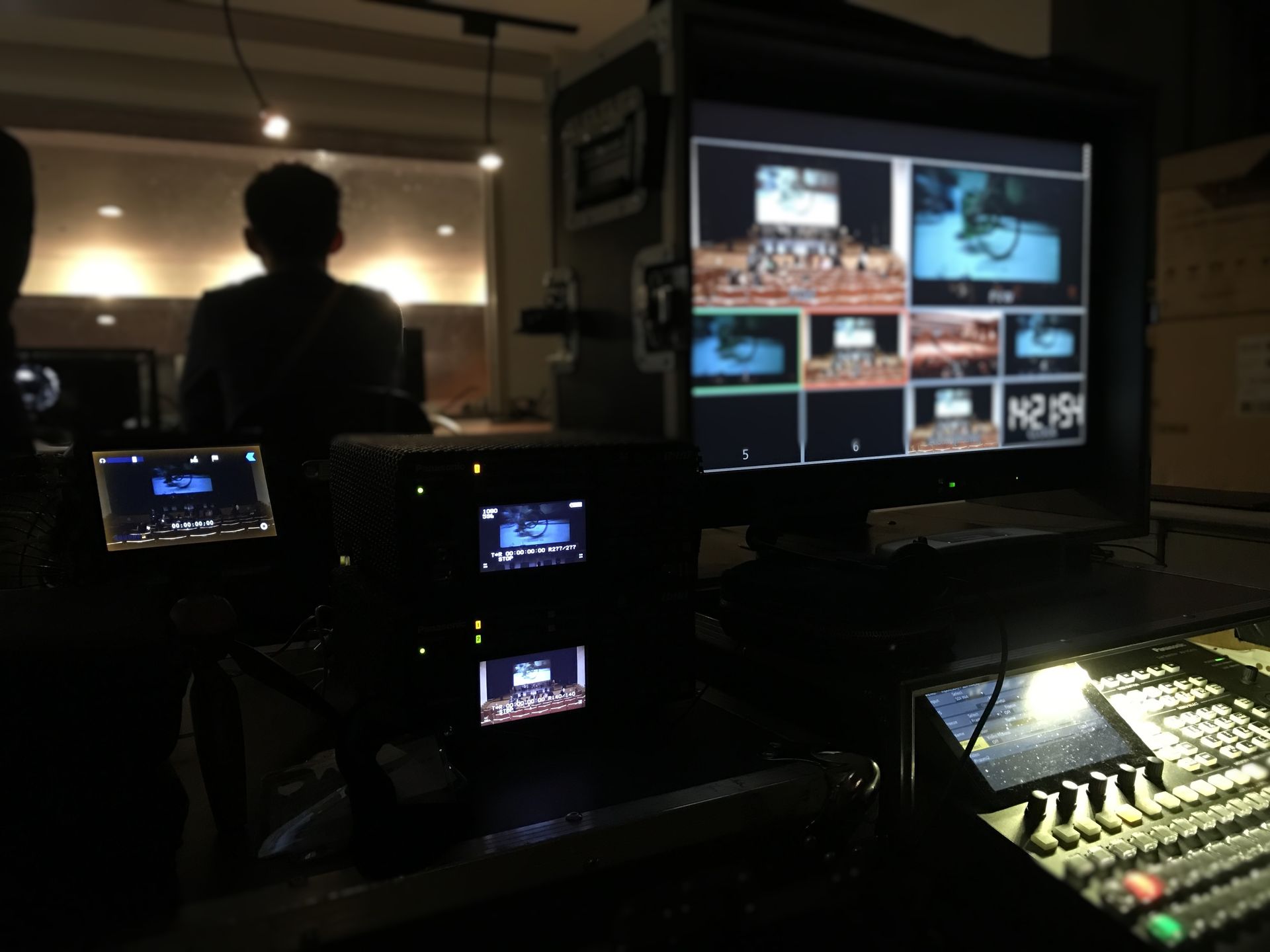 廣播控制室，控制台旁的人員，多個視訊顯示器顯示即時畫面和控制面板。