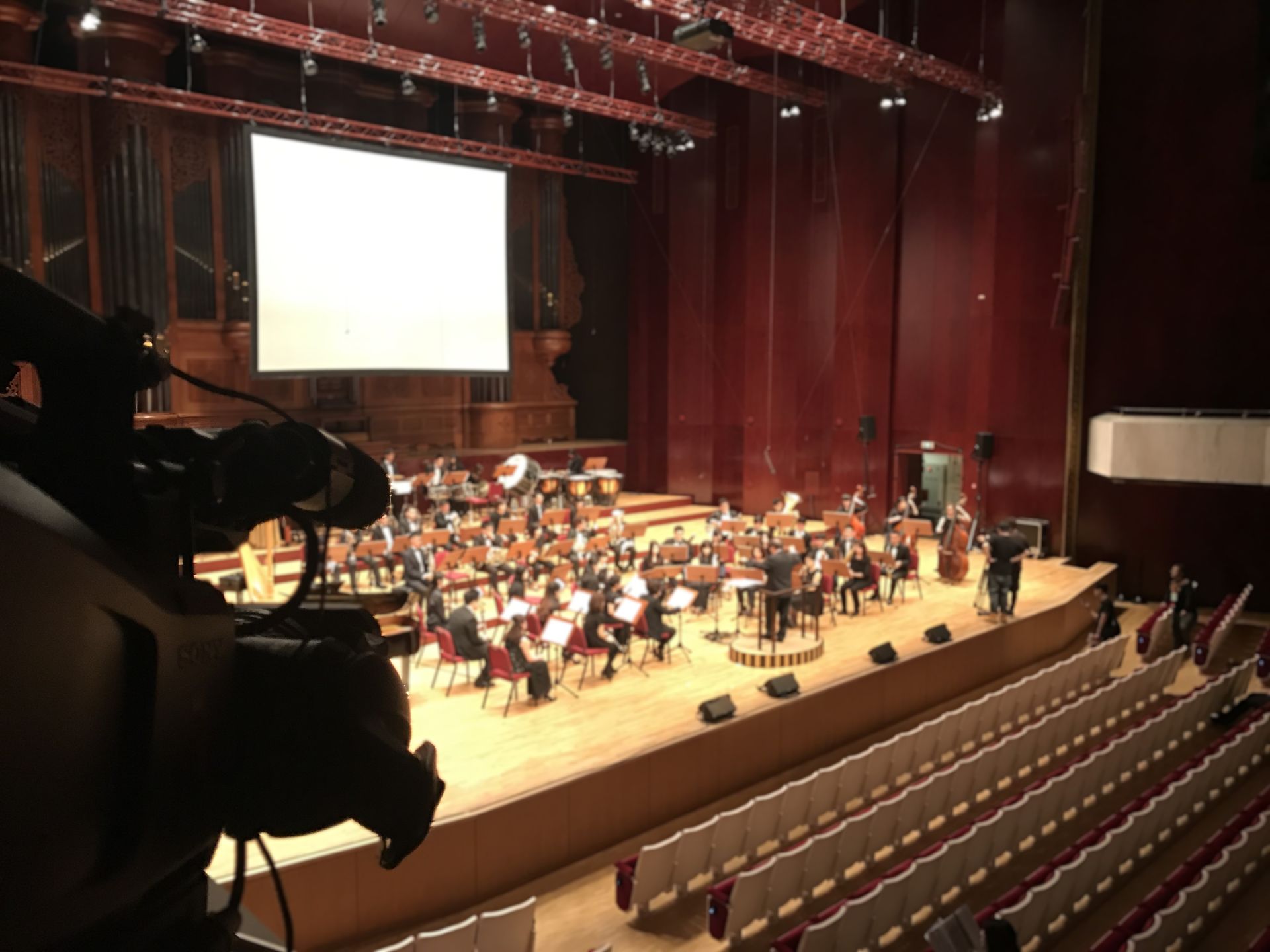 一支管弦樂團在舞台上表演，前景中放置著一台攝影機。紅色的牆壁和空蕩蕩的座位。