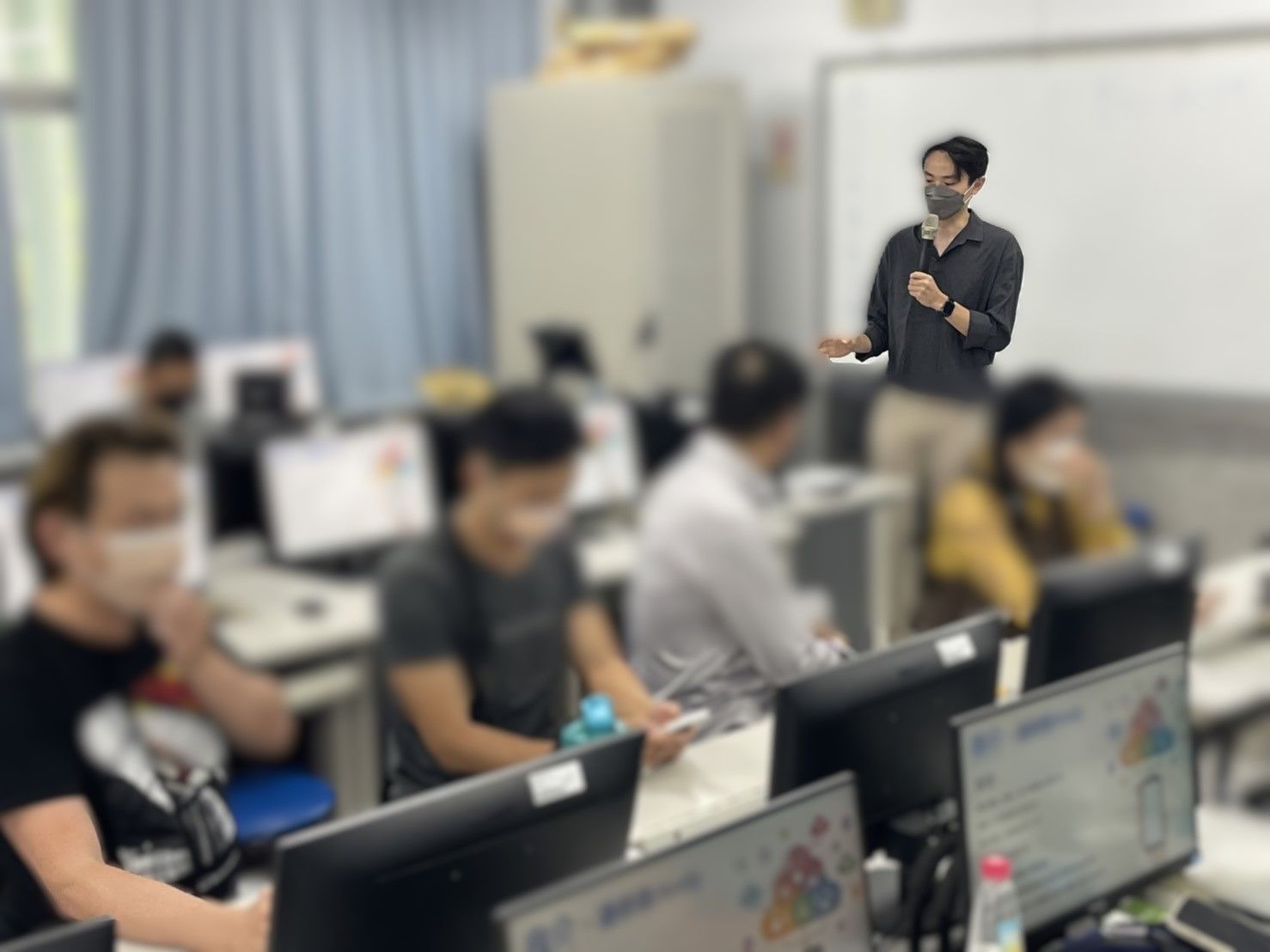 一位戴著面具的老師在電腦實驗室裡指著白板給學生講課。