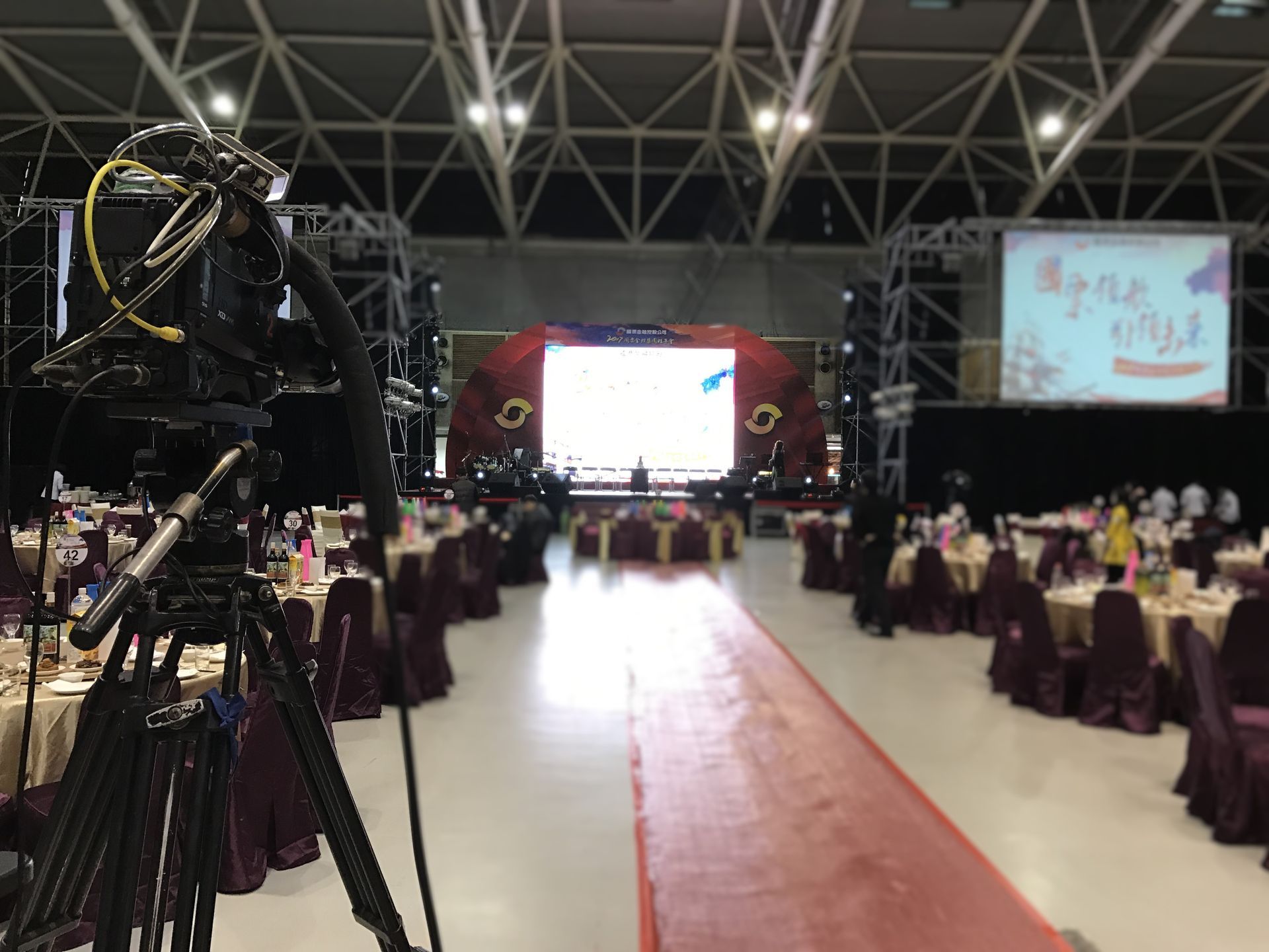 大廳裡，三腳架上的攝影機拍攝活動舞台，宴會桌擺放整齊。