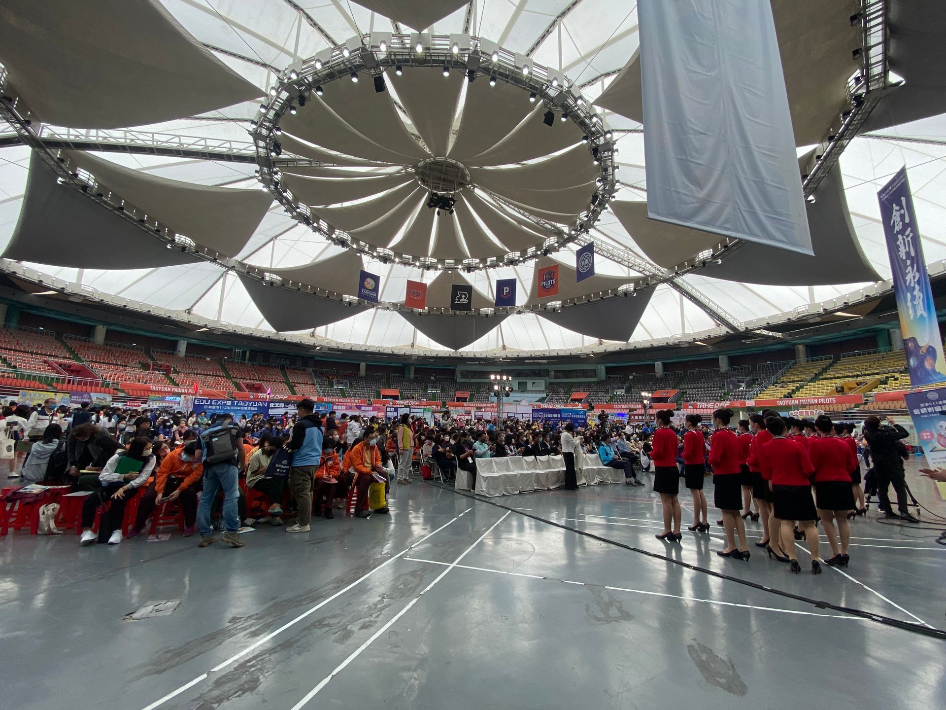 大型體育場內，人們正在觀看一場活動，體育場的頂棚可伸縮。紅色和橙色的座位清晰可見。
