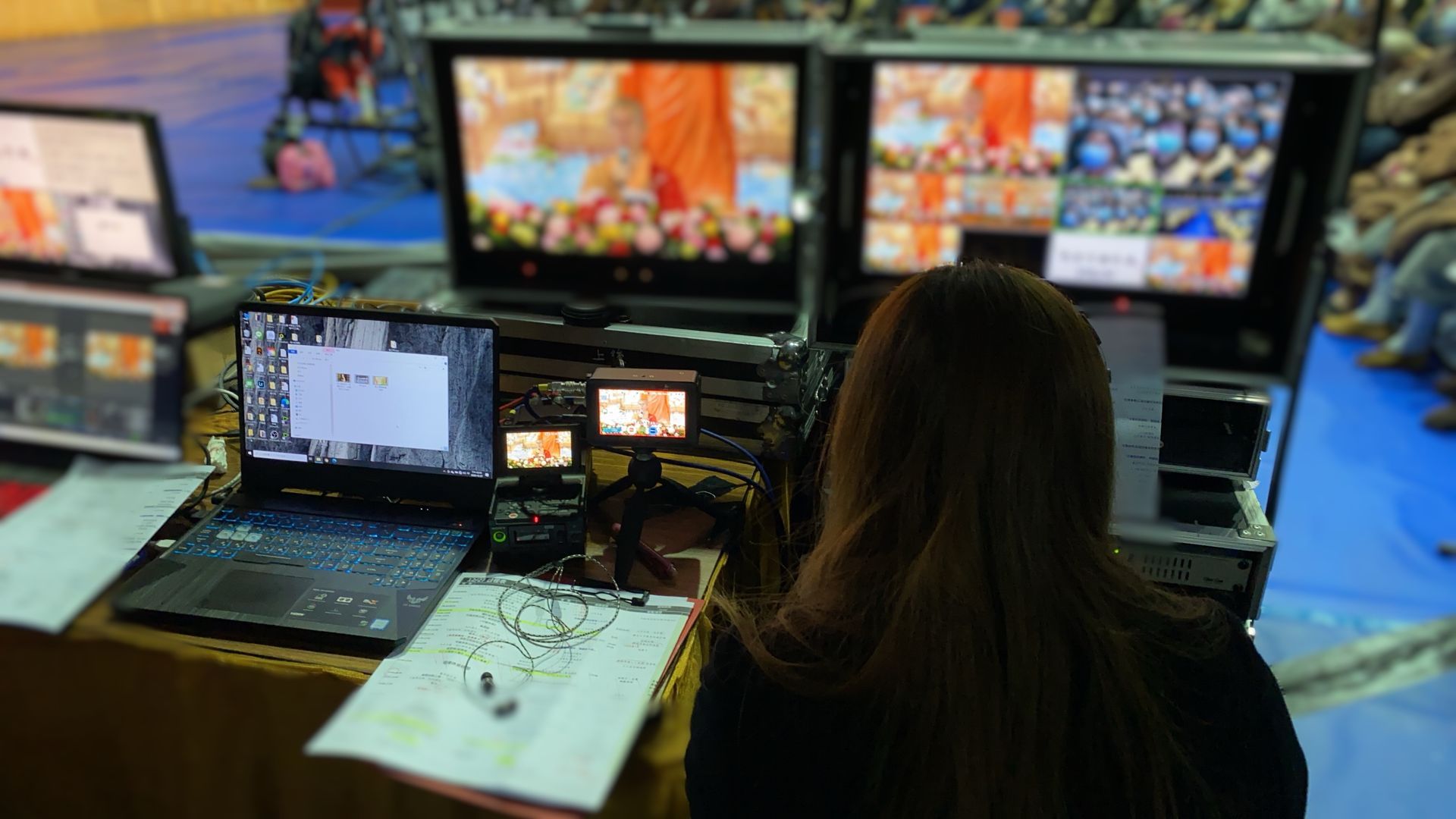 影片製作站的女人，監視器顯示現場鏡頭，桌上有圖表。