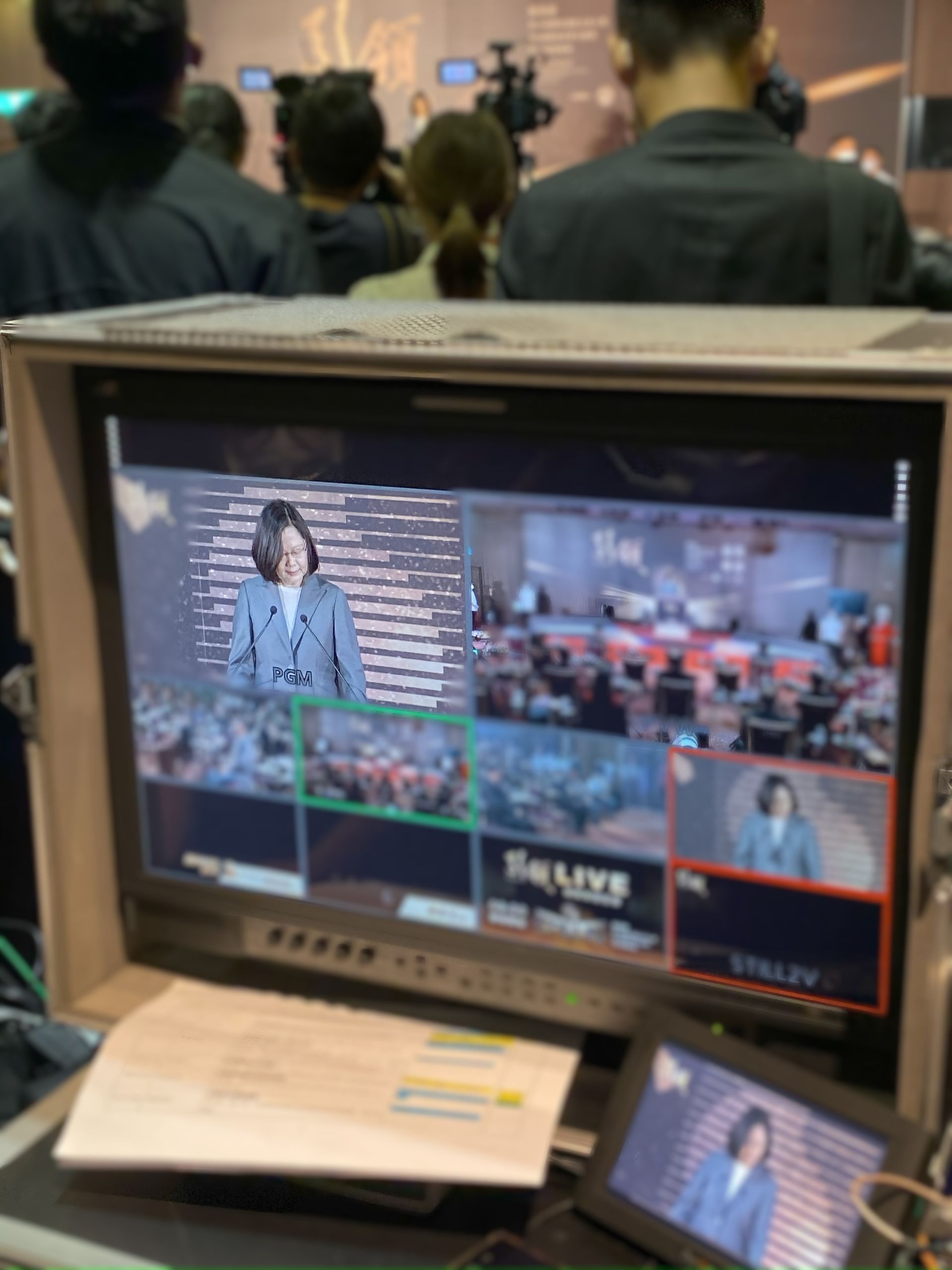 監視器正在直播一位女士在講台上講話的場景，前景是一間控制室。