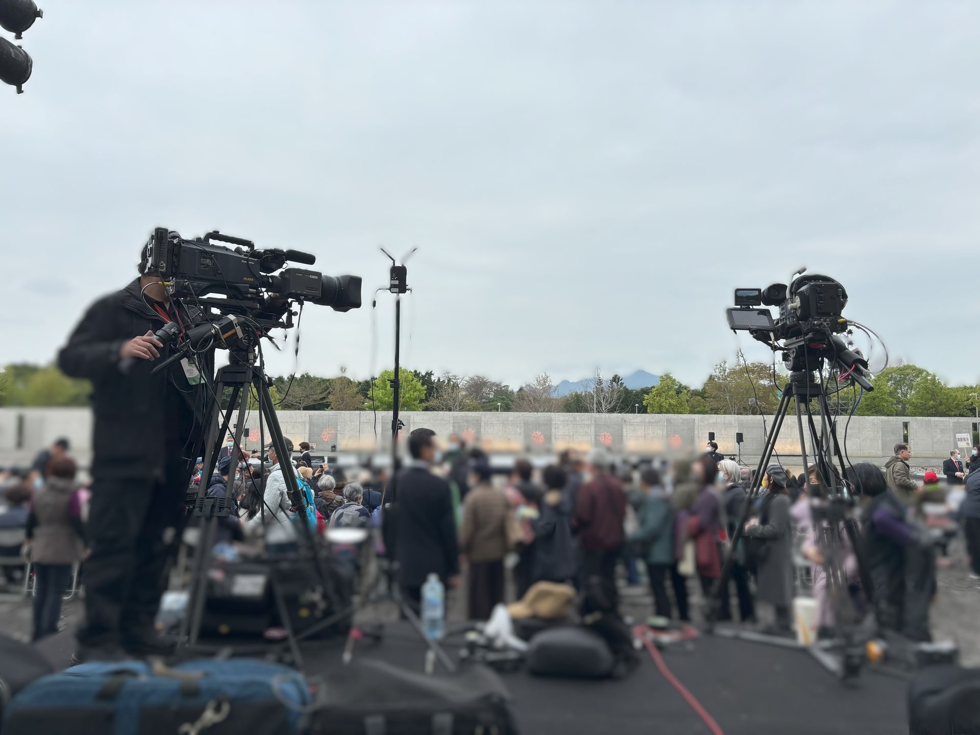 兩台安裝在三腳架上的廣播攝影機正在拍攝戶外場景中的人群，頭頂上是多雲的天空。