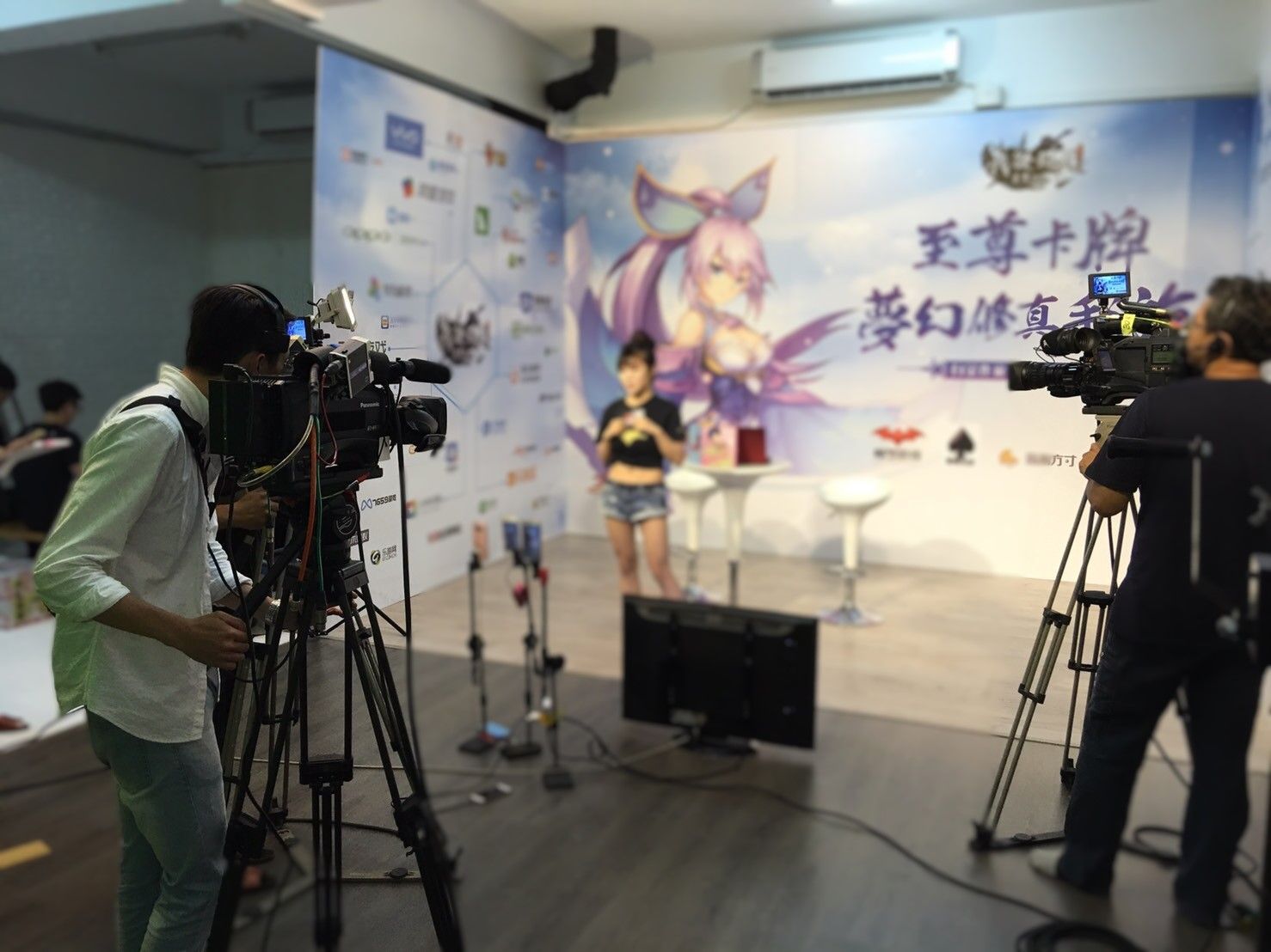 一位身穿短褲的女子站在片場，對著兩台攝影機講話。工作人員在攝影棚裡操作著攝影機。