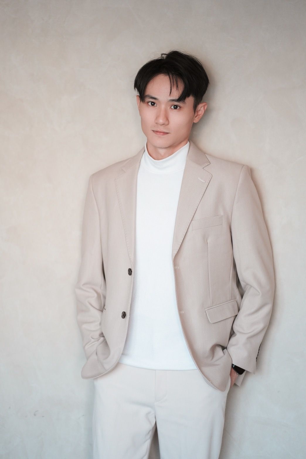 一名有著深色頭髮的年輕人，身穿奶油色外套、白色襯衫和淺色褲子，靠在牆上。