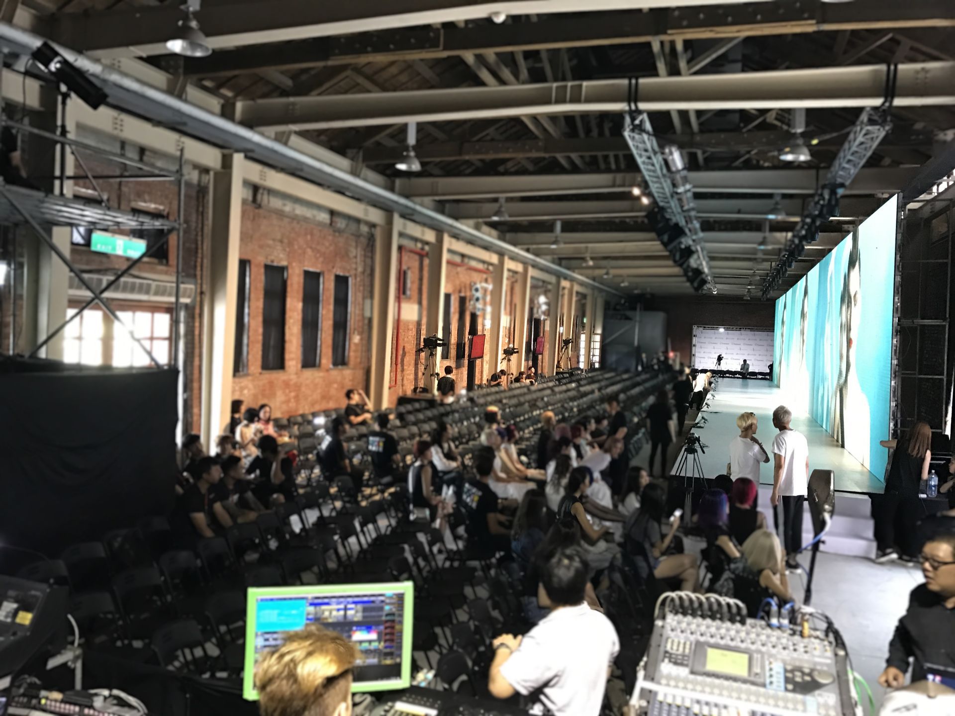 時裝秀：賓客入座，觀賞模特兒走秀和大螢幕。磚造建築內部，頭頂燈光。