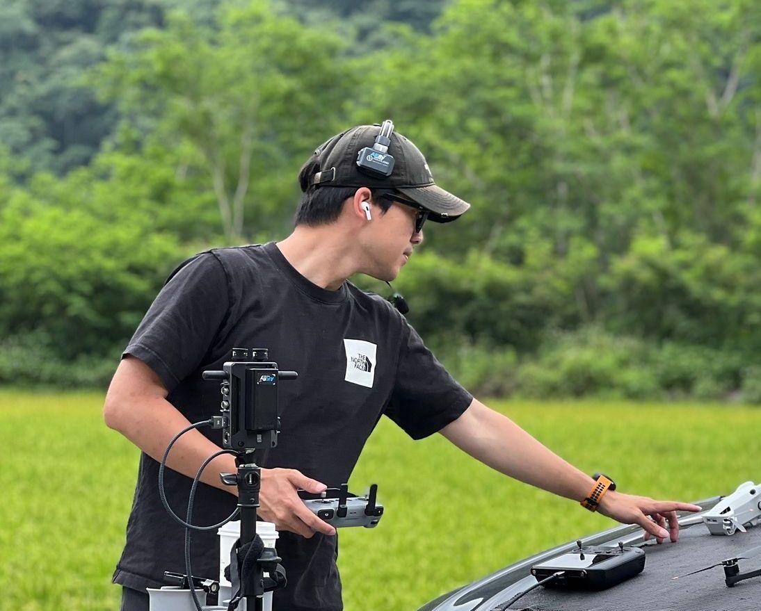 身穿黑色襯衫、頭戴黑色帽子的男子在戶外操作無人機,指著無人機表面。背景是綠色的田野和樹木。