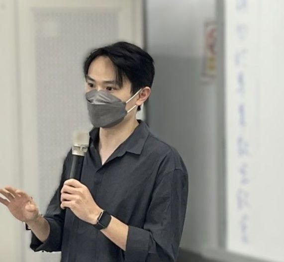 一名男子身穿灰色襯衫，戴著面具，手裡拿著麥克風，在白板前做手勢。