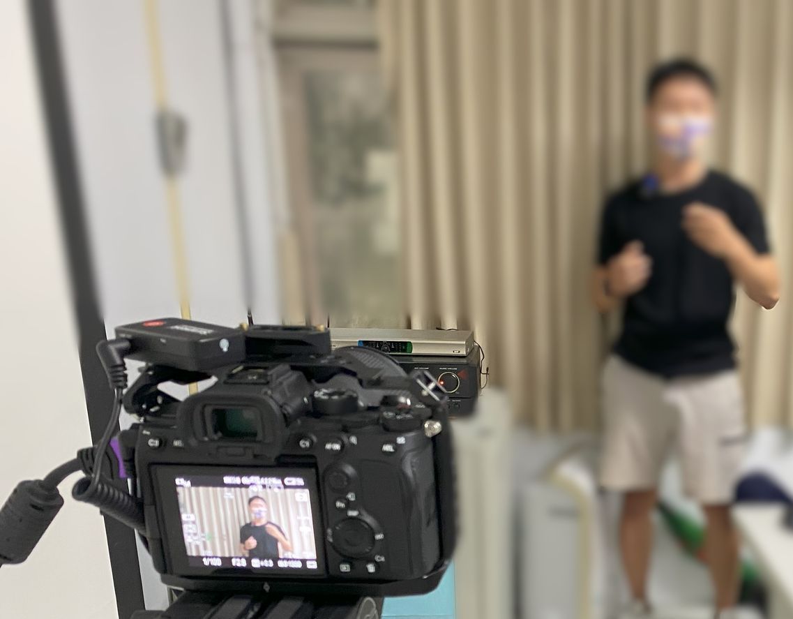 攝影機拍攝到一名身穿短褲和黑色襯衫的男子,他站在掛著米色窗簾的窗戶前做手勢。