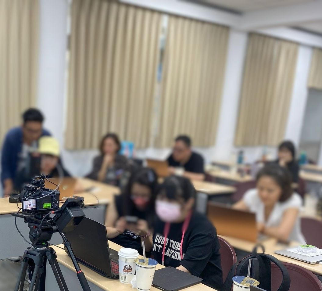 教室裡,人們拿著筆記型電腦。三腳架上架著一台攝影機,準備記錄他們。