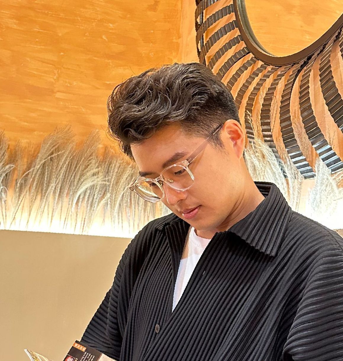 一名男子戴眼鏡,低頭看著前方,身穿黑色領襯衫。背景是淺色的木頭。