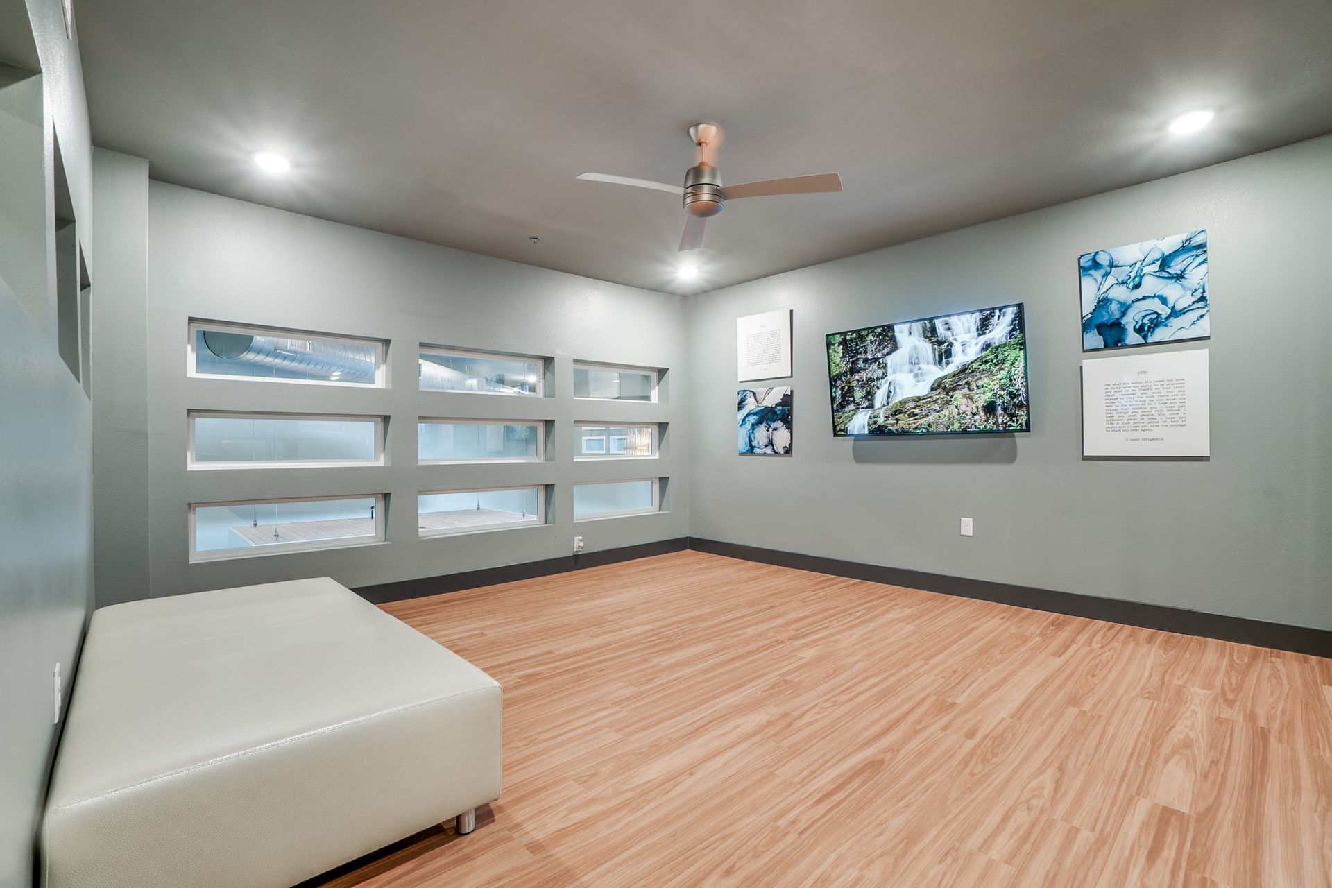 Interior community lounge with wood-like flooring, gray walls, a ceiling fan, bench seating, and wall art.