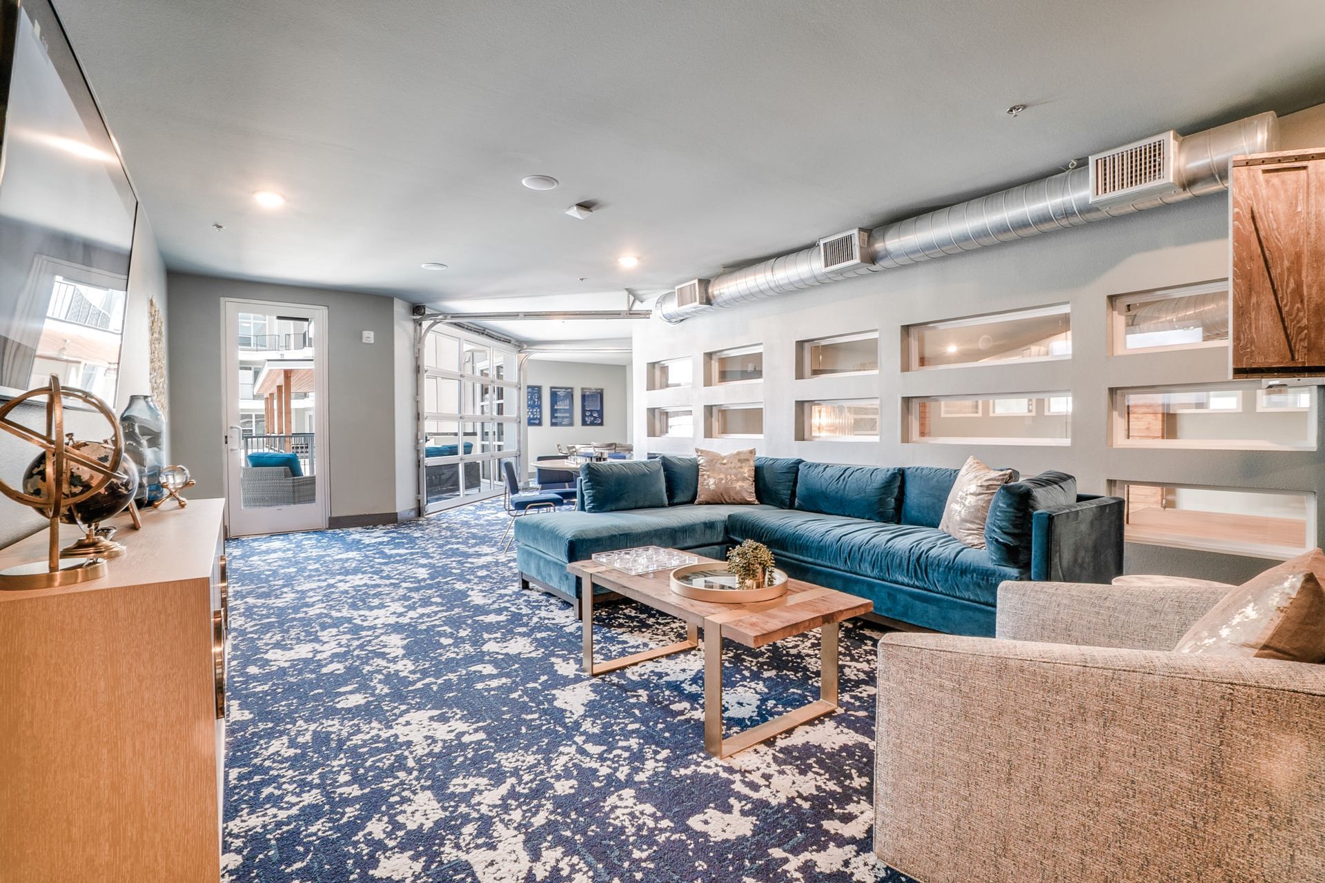 Indoor community lounge with a large blue sectional sofa, coffee table, and exposed ductwork.