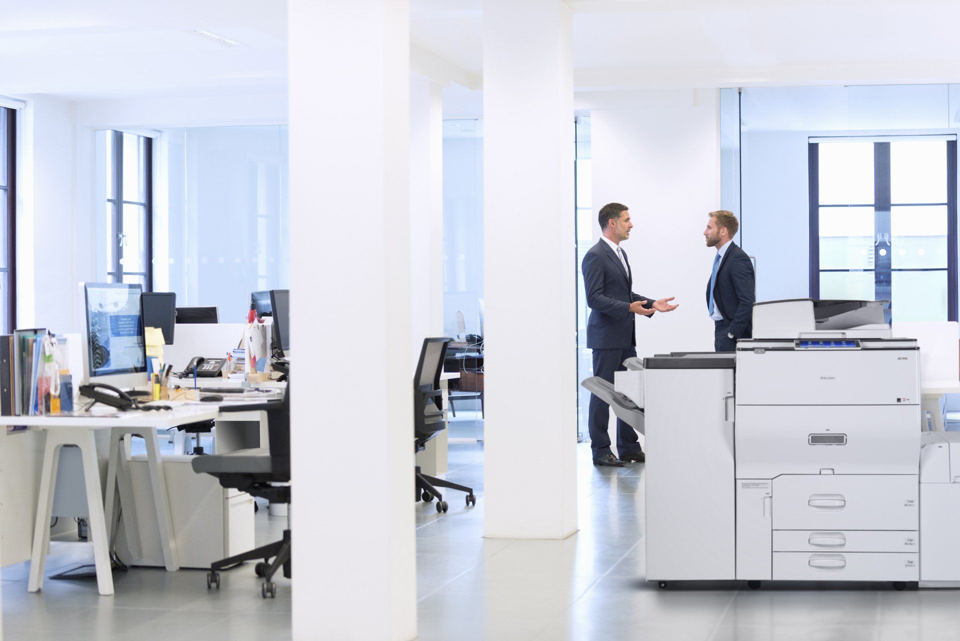 Two men are standing next to each other in an office talking to each other.